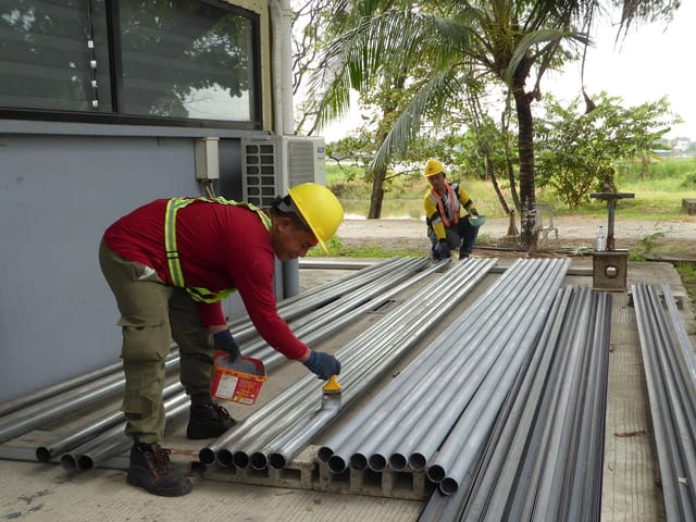 Workers preparing steel pipework for coating at an outdoor industrial site