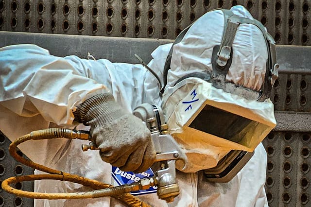 Worker in a protective suit holding a blast nozzle and hose against a steel surface