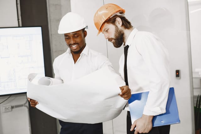 Two engineers in hard hats reviewing a plant drawing in front of a screen